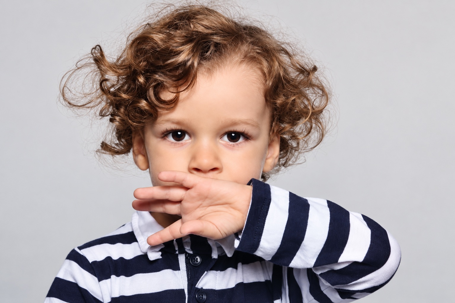 A curly-haired child in a striped shirt with a hand near their mouth.